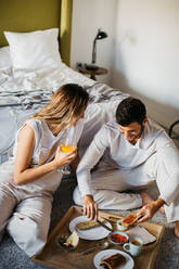 Von oben auf das glückliche junge Paar, das in der Nähe des Bettes zusammensitzt und kommuniziert, während es das Frühstück mit Gebäck und Getränken während der Flitterwochen genießt - ADSF38018