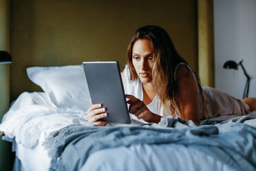 Young woman touching screen of tablet while lying on bed in light modern apartment - ADSF38023