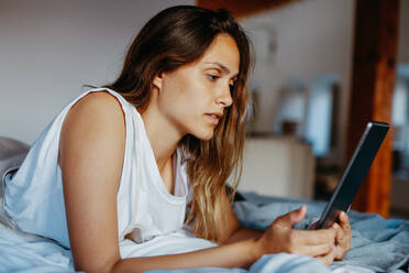 Seitenansicht einer jungen Frau, die den Bildschirm eines Tablets berührt, während sie auf dem Bett in einer hellen, modernen Wohnung liegt - ADSF38024