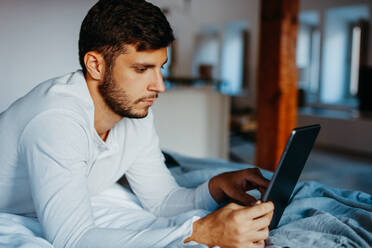 Seitenansicht eines jungen, bärtigen Mannes, der den Bildschirm eines Tablets berührt, während er auf dem Bett in einer hellen, modernen Wohnung liegt - ADSF38025