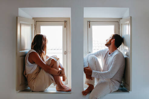 Side view of barefoot couple in domestic cloth sitting on nearby windowsills and looking out window on sunny day at home - ADSF38027
