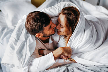 Von oben auf ein glückliches junges Paar, das sich zärtlich umarmt, während es am Morgen unter einer warmen Decke im Bett liegt - ADSF38029
