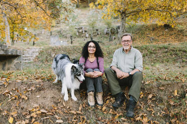 Full length cheerful mature couple in casual wear sitting on ground near cute Australian Shepherd dog in autumn park and looking at camera with smiles - ADSF38035