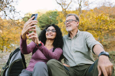 Zufriedenes älteres Paar in legerer Kleidung und mit Brille, das ein Selfie mit einem modernen Smartphone macht, während es zusammen auf einem grasbewachsenen Boden im Herbstpark sitzt - ADSF38040