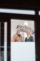 Fröhliche ältere Frau lächelt und hält den Schirm über einen Mann mit medizinischer Maske, während sie auf der Terrasse eines Anwesens steht - ADSF38049