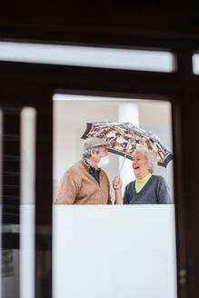 Cheerful elderly woman smiling and keeping umbrella over man in medical mask while standing on terrace of estate - ADSF38049