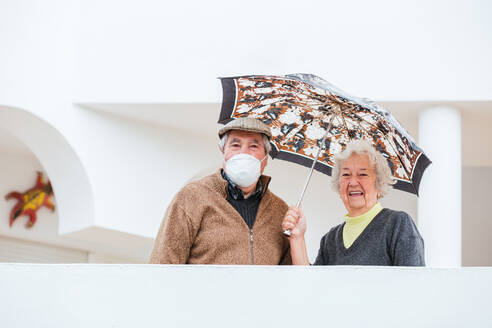 Cheerful elderly woman smiling and keeping umbrella over man in medical mask while standing on terrace of estate - ADSF38050