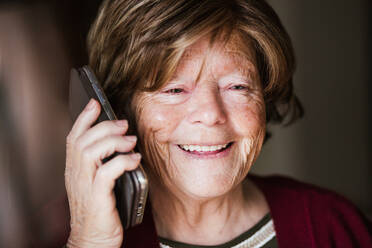 Ältere Dame mit faltigem Gesicht, die wegschaut und einen Telefonanruf entgegennimmt, während sie zu Hause ist - ADSF38051