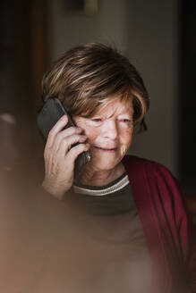Ältere Dame mit faltigem Gesicht, die wegschaut und einen Telefonanruf entgegennimmt, während sie zu Hause ist - ADSF38052