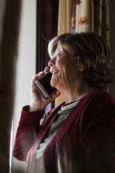 Seitenansicht einer glücklichen älteren Dame, die einen Telefonanruf entgegennimmt, während sie zu Hause ist - ADSF38055