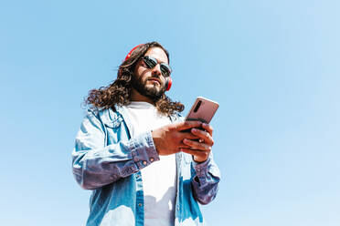 From below bearded Latin American male in modern sunglasses browsing on cellphone and wireless headphones on blue background on sunny day - ADSF38124