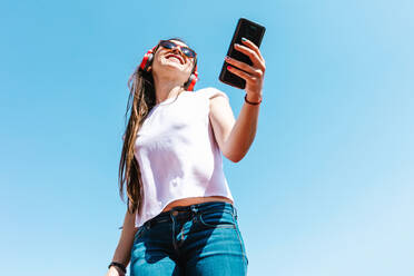 Von unten lateinamerikanische Frau in weißem T-Shirt und moderne Sonnenbrille mit Handy und drahtlose Kopfhörer auf blauem Hintergrund auf sonnigen Tag - ADSF38125