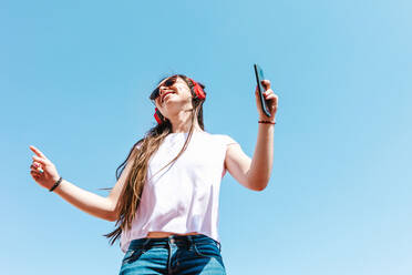 Von unten lateinamerikanische Frau in weißem T-Shirt und moderne Sonnenbrille mit Handy und drahtlose Kopfhörer auf blauem Hintergrund auf sonnigen Tag - ADSF38126