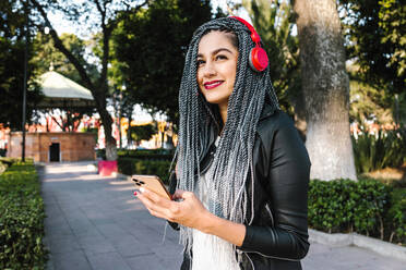 Cheerful ethnic female with Afro braids in modern headset browsing on smartphone in town looking away - ADSF38135