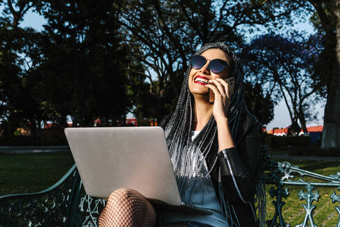 Positive lateinamerikanische Frau in modischer Kleidung und Sonnenbrille mit Netbook, die auf einer Bank im Sonnenlicht sitzt und mit ihrem Handy telefoniert - ADSF38142