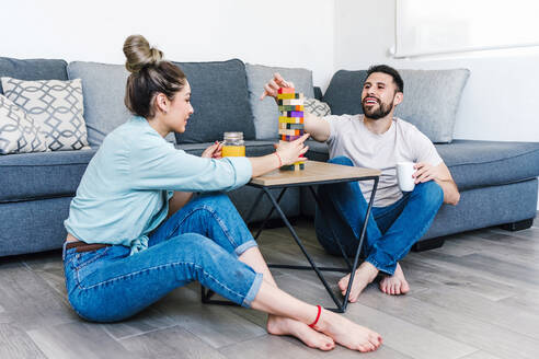 Positives lateinisches Paar, das am Tisch ein Turmspiel mit Holzklötzen spielt, während es sich am Wochenende zu Hause unterhält - ADSF38153