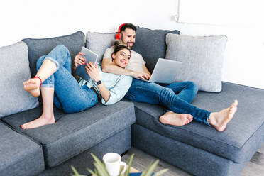 Content Latin couple of freelancers using tablet and laptop while sitting on sofa and working on remote project together from home - ADSF38156