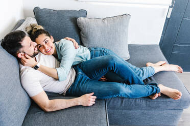 High angle of tender Latin couple reading book together while embracing and lying on couch in living room at home - ADSF38158