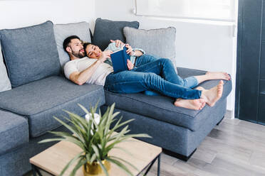 Zartes lateinisches Paar, das zusammen ein Buch liest, während es sich umarmt und auf der Couch im Wohnzimmer zu Hause liegt - ADSF38159