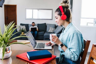 Seitenansicht einer lateinamerikanischen Freiberuflerin mit Kopfhörern und einer Tasse Kaffee, die auf einem Netbook surft, während sie an einem Projekt am Tisch zu Hause mit einem unscharfen Freund arbeitet - ADSF38161