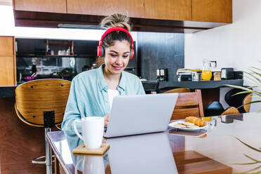 Lateinische Freiberuflerin mit Kopfhörern und einer Tasse Kaffee, die auf ihrem Netbook surft, während sie an einem Projekt am Tisch zu Hause arbeitet - ADSF38164