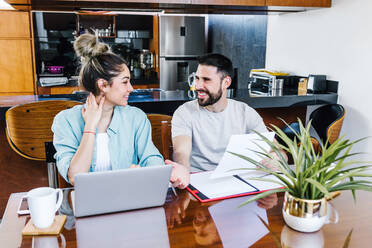 Content ethnic couple of freelancers browsing laptop and reading documents while working on project together in kitchen at home looking at each other - ADSF38168