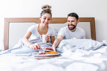 Low angle of happy ethnic couple chilling in bed and enjoying tasty breakfast in morning at home - ADSF38169