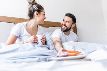 Low angle of happy ethnic couple chilling in bed and enjoying tasty breakfast in morning at home looking at each other - ADSF38170