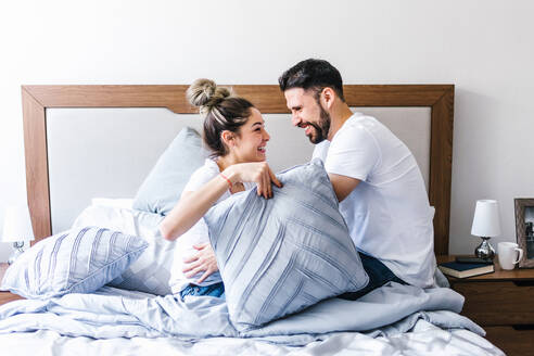 Seitenansicht eines sanften lateinischen Mannes, der seine Freundin umarmt, während er sich mit einem Kissen auf einem weichen Bett im Schlafzimmer versteckt und sich gegenseitig ansieht - ADSF38171