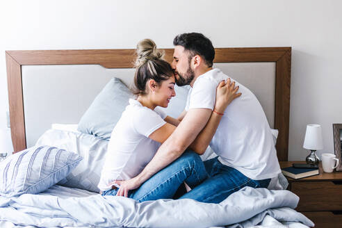 Seitenansicht eines sanften lateinischen Mannes, der seine Freundin auf die Stirn küsst, während er sich auf einem weichen Bett im Schlafzimmer umarmt - ADSF38172