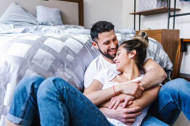 Liebendes ethnisches Paar, das sich lächelnd umarmt, während es im Schlafzimmer auf dem Boden sitzt und sich gegenseitig anschaut - ADSF38176