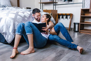 Positive ethnic couple hugging and reading book together while sitting of floor in bedroom and looking at each other - ADSF38177