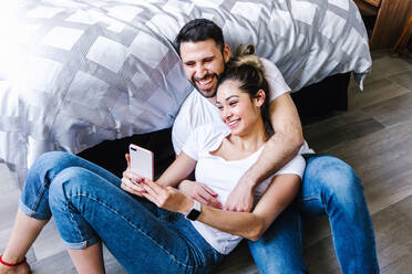 Fröhliches lateinamerikanisches Paar, das sich umarmt und ein Selfie mit dem Handy macht, während es sich zu Hause amüsiert - ADSF38180