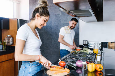 Begeistertes lateinamerikanisches Paar, das frisches reifes Gemüse schneidet und einen gesunden Salat für das Mittagessen in der Küche zu Hause zubereitet und sich dabei gegenseitig ansieht - ADSF38181