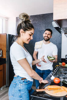 Begeistertes lateinamerikanisches Paar, das frisches reifes Gemüse schneidet und einen gesunden Salat für das Mittagessen in der Küche zu Hause zubereitet und sich dabei gegenseitig ansieht - ADSF38182