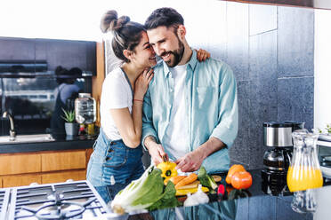 Delighted Latin couple embracing while cutting fresh ripe vegetables and preparing healthy salad for lunch in kitchen at home - ADSF38184