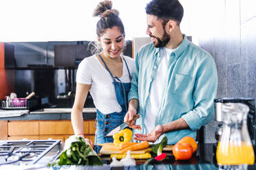 Vergnügtes lateinamerikanisches Paar, das sich umarmt, während es frisches reifes Gemüse schneidet und einen gesunden Salat für das Mittagessen in der Küche zu Hause zubereitet - ADSF38185