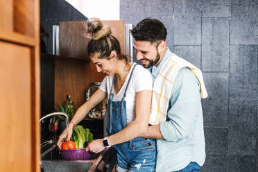 Side view of delighted ethnic woman washing fresh vegetables with boyfriend embracing her while preparing lunch at home - ADSF38192