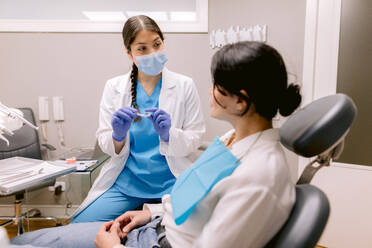 Weiblicher Kieferorthopäde in Uniform und steriler Maske mit Zahnspange im Gespräch mit einer Frau während einer zahnärztlichen Beratung, während sie sich gegenseitig im Krankenhaus ansehen - ADSF38213