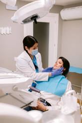 Female orthodontist in uniform and sterile mask checking up jaw of woman on dental chair in hospital - ADSF38218