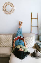 Relaxed young woman reading a book at home while sitting on couch - ADSF38233
