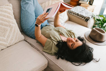 Entspannte junge Frau, die zu Hause auf der Couch sitzend ein Buch liest - ADSF38234