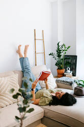 Relaxed young woman reading a book at home while sitting on couch - ADSF38235