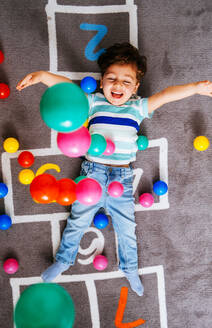 Von oben aufgeregter Junge in Freizeitkleidung, der lachend bunte Bälle in die Luft wirft, während er auf dem Boden liegt und mit Zahlen hüpft und zu Hause spielt - ADSF38269