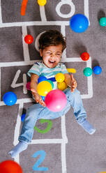 Von oben aufgeregter Junge in Freizeitkleidung, der lachend bunte Bälle in die Luft wirft, während er auf dem Boden liegt und mit Zahlen hüpft und zu Hause spielt - ADSF38270