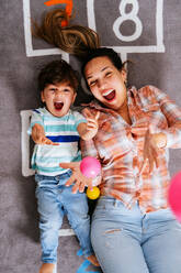 From above delighted woman and little boy screaming and throwing colorful balls up while lying on nursery floor together - ADSF38271