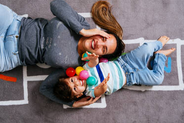 From above delighted woman and little boy screaming and throwing colorful balls up while lying on nursery floor together - ADSF38273