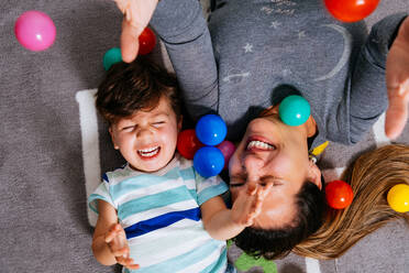 From above delighted woman and little boy screaming and throwing colorful balls up while lying on nursery floor together - ADSF38274