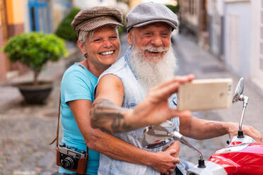 Seitenansicht eines erwachsenen Paares, das ein Selfie mit seinem Smartphone macht, während es mit dem Motorrad durch die Stadt fährt - ADSF38280