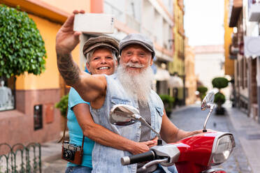 Seitenansicht eines älteren Paares, das ein Selfie mit seinem Smartphone macht, während es mit dem Motorrad durch die Stadt fährt - ADSF38281